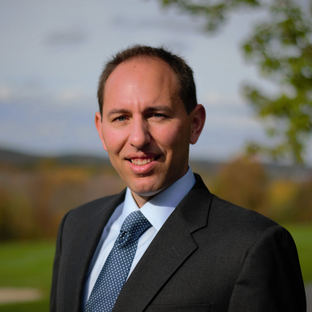 This photo features Bill Suzor, proud parent of Lila, his 11-year-old daughter affected by ECHS1 Deficiency and Director of Cure Mito, an organization dedicated to finding a cure for mitochondrial diseases. He is smiling, wearing a dark suit jacket, light blue shirt, and patterned tie, with short brown hair. This image highlights the leadership and personal connection driving Cure Mito's mission to improve the lives of those affected by these conditions.